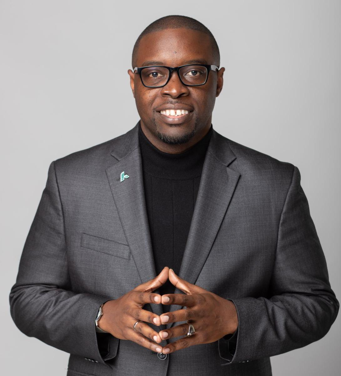 Dr. Jermaine Whirl, a smiling African American male, wearing a black suit with a white collared shirt, a brown tie with gold dots, and black rimmed glasses. A round gold pin is on his left lapel.